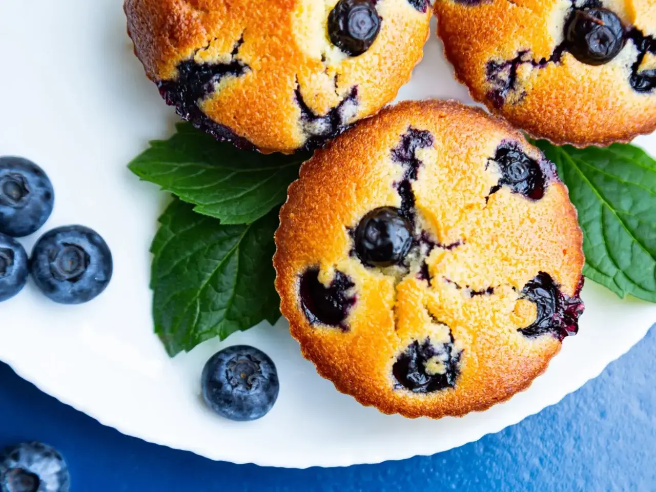 Jak udekorować muffinki? Puszyste babeczki z jagodami, świeże borówki i zielone listki mięty na białym talerzu.