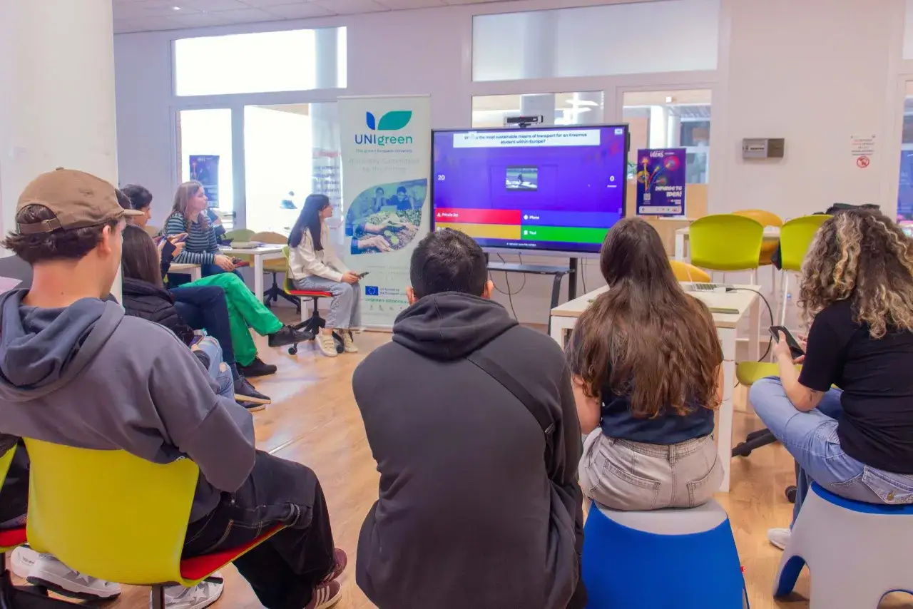 J&oacute;venes discutiendo sobre transporte sostenible para becas Erasmus, mientras un juego interactivo se muestra en pantalla.