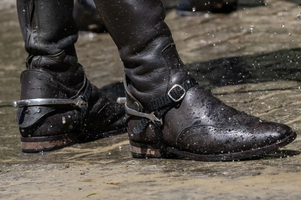 Schwarze Lederstiefel mit Sporen, die im Wasser stehen. Die Sporen glänzen und die Stiefel sind nass.