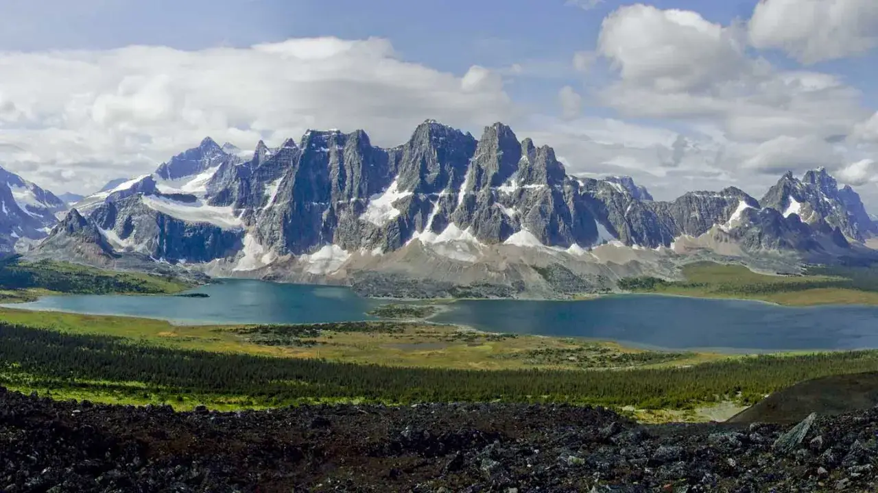 Majestic peaks of the Tonquin Valley rise above a serene blue lake, with lush green meadows and dark, rocky foreground.