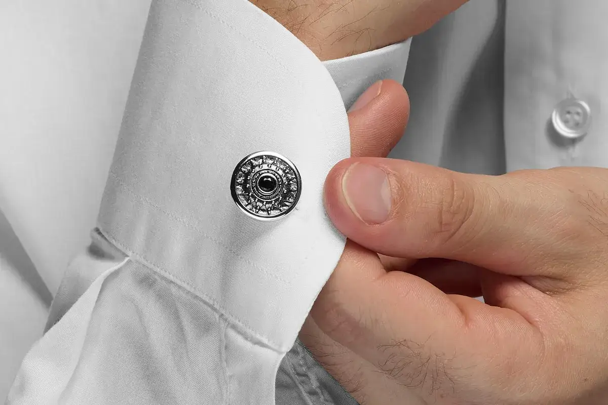 Dettaglio su come si mettono i gemelli: una mano sistema un gemello rotondo con decorazioni intricate su un polsino di camicia bianca.