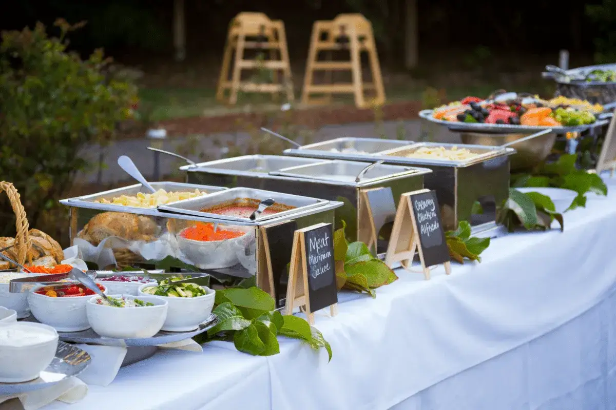 Buffet de mariage sans traiteur : plats chauds, salades et pains disposés sur une longue table blanche, décorée de feuilles vertes.