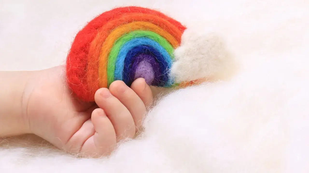 Dłoń niemowlaka trzyma tęczowe dziecko – małą, filcową tęczę z chmurką.