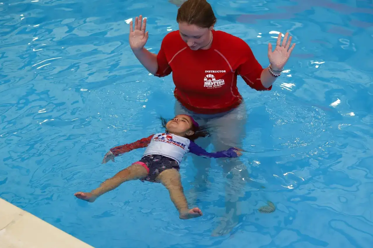 A swim instructor helps a young child float on their back, learning to safe t swim. The child looks relaxed in the blue water.