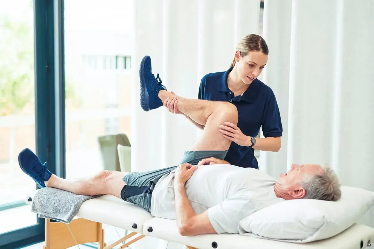 Physiotherapeutin hilft älterem Mann bei der Beinübung. Aktive Behandlung kann die Lebensqualität bei Arthrose verbessern.