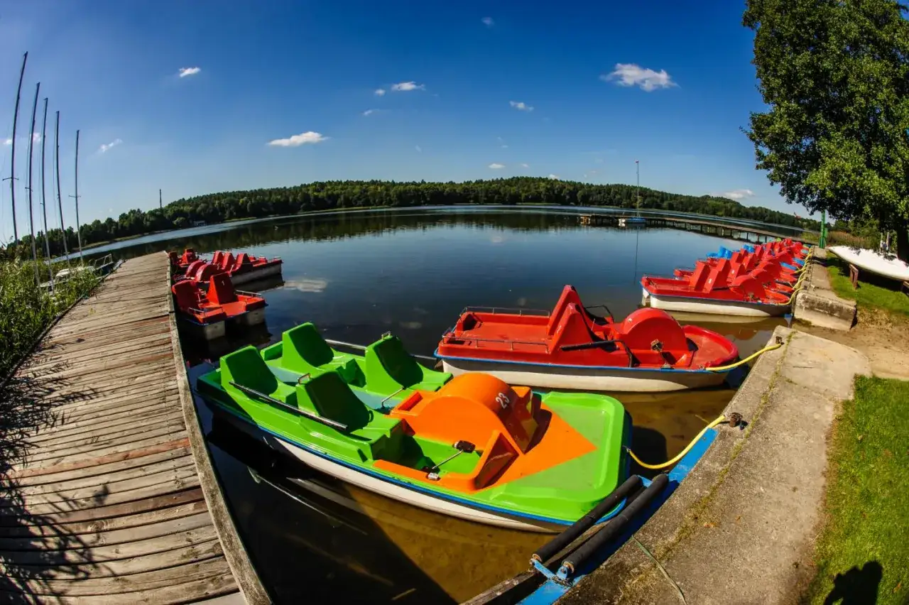 Kolorowe rowerki wodne czekają na plaży nad jeziorem Kortowskim. Drewniany pomost i spokojna tafla wody zapraszają do relaksu.