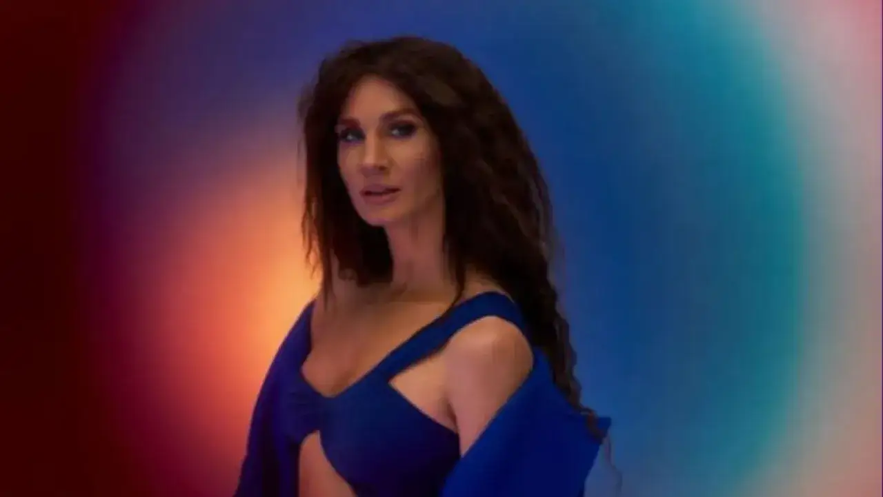 Sylwia Grzeszczak w niebieskiej kreacji, z burzą ciemnych włosów, na tle tęczowego gradientu. Jej wiek jest nieistotny wobec jej talentu.