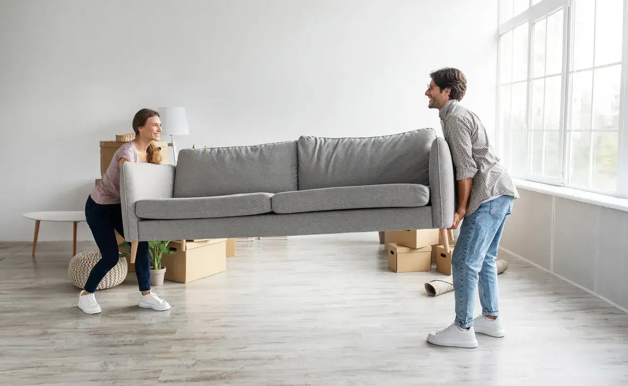 Pareja moviendo un sofá gris en una habitación vacía, preparándose para invertir en inmuebles para alquilar.