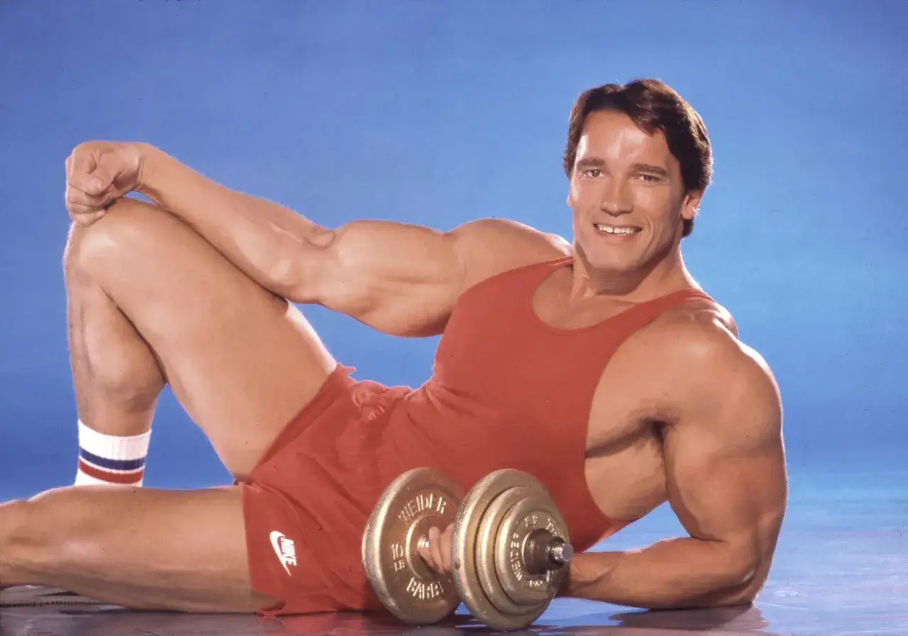 Arnold Schwarzenegger, znany z imponującego wzrostu i wagi, prezentuje swoją muskulaturę w czerwonym stroju sportowym, trzymając hantle.