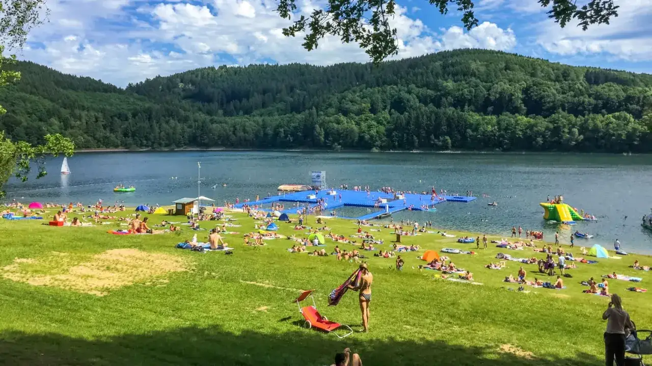 Sonniger Tag am Badesee OWL mit vielen Menschen, die sich auf der Wiese sonnen und im Wasser planschen. Eine blaue schwimmende Plattform und eine Wasserrutsche sorgen für Spaß.