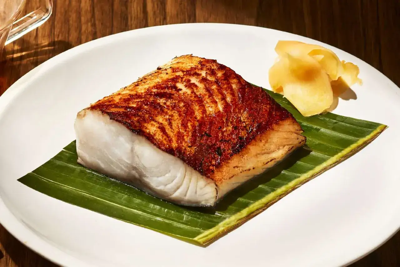 A perfectly grilled nobu miso black cod fillet rests on a banana leaf, served with pickled ginger on a white plate.