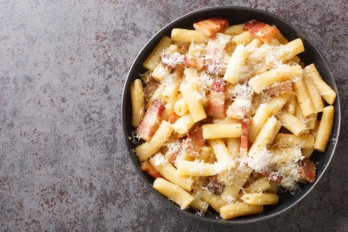 Rigatoni alla gricia fumanti, con guanciale croccante e pecorino grattugiato, serviti in una ciotola scura.
