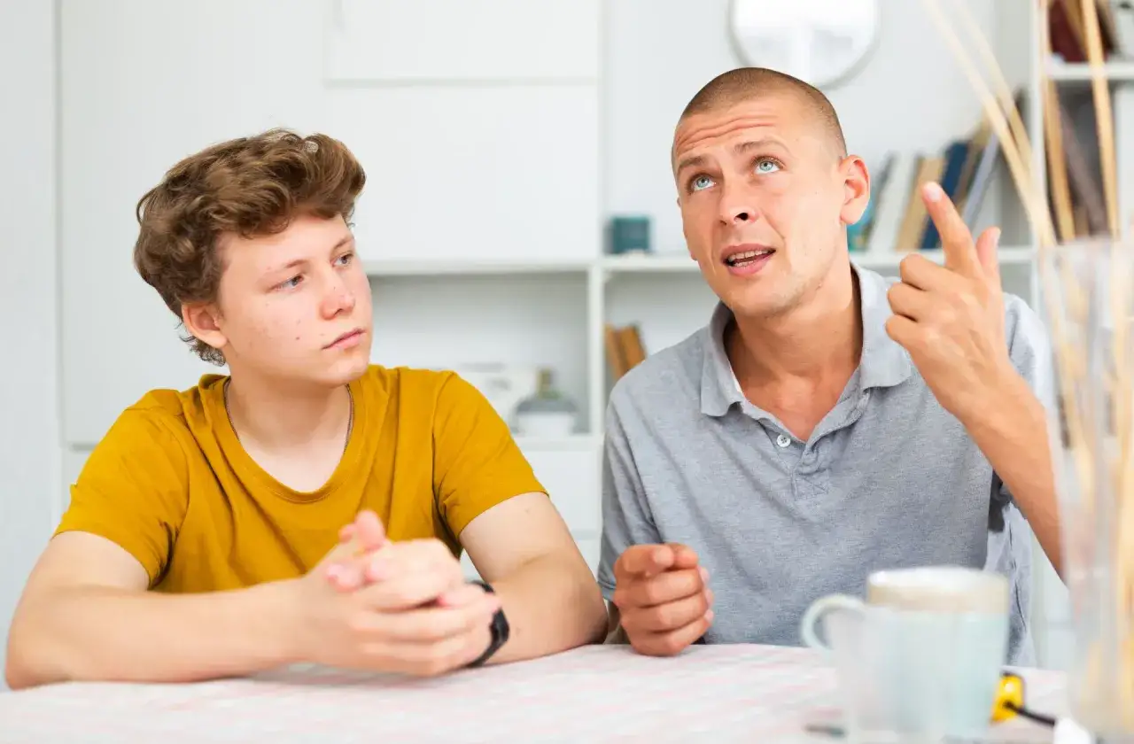 Ojciec tłumaczy synowi, jak rozmawiać z nastolatkiem, gestykulując i patrząc w górę. Chłopak słucha uważnie.