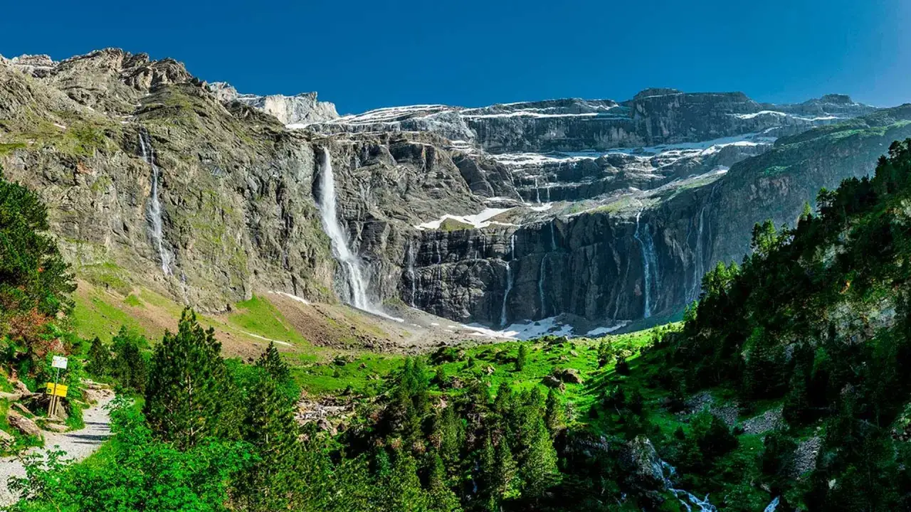 Cascadas impresionantes caen por acantilados rocosos en el **lac de Gaube**, rodeado de verdes praderas y pinos.