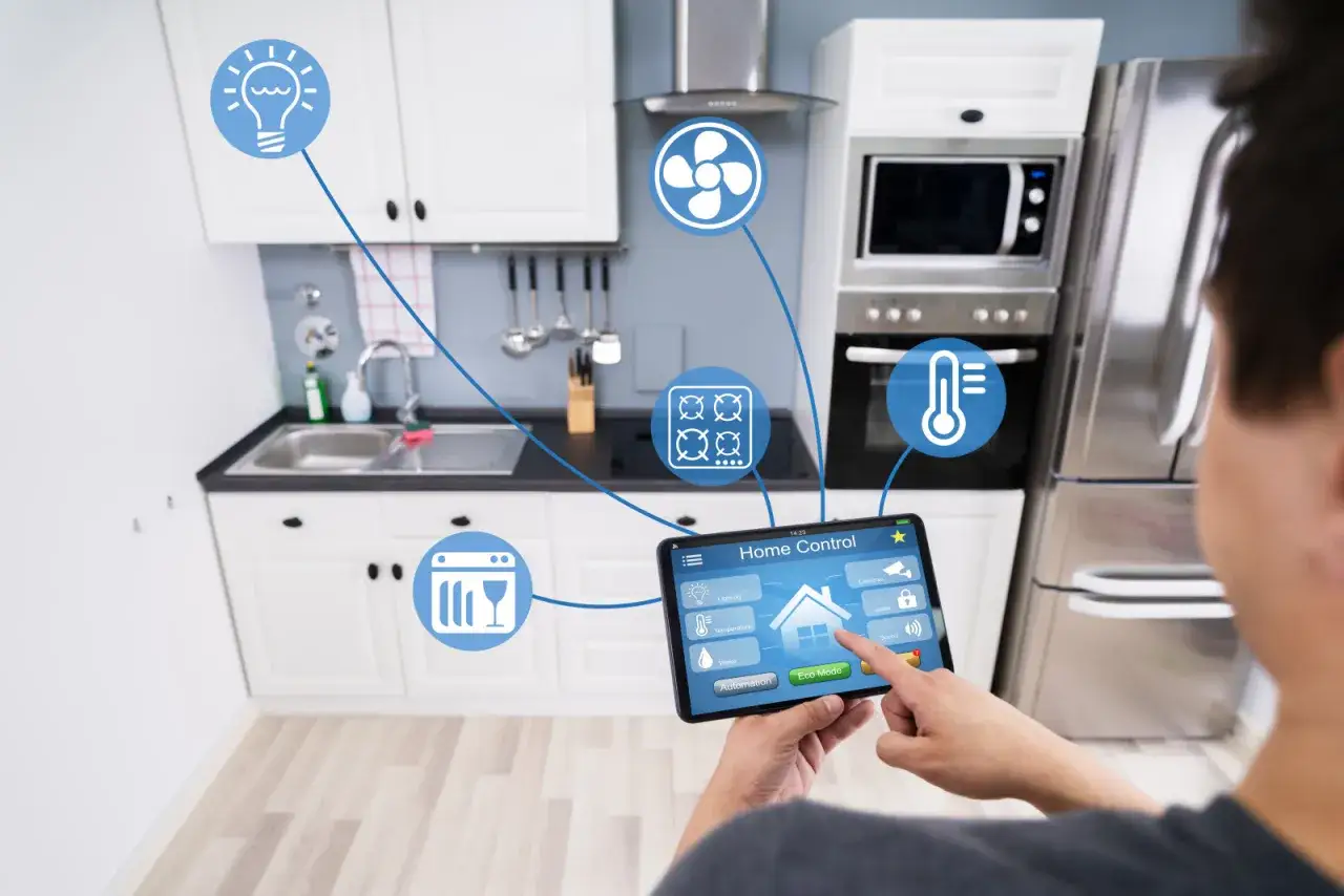 Mężczyzna steruje urządzeniami w kuchni za pomocą tabletu, realizując wizję smartlife. Widoczne ikony: żarówka, wentylator, płyta grzewcza, zmywarka, termometr.