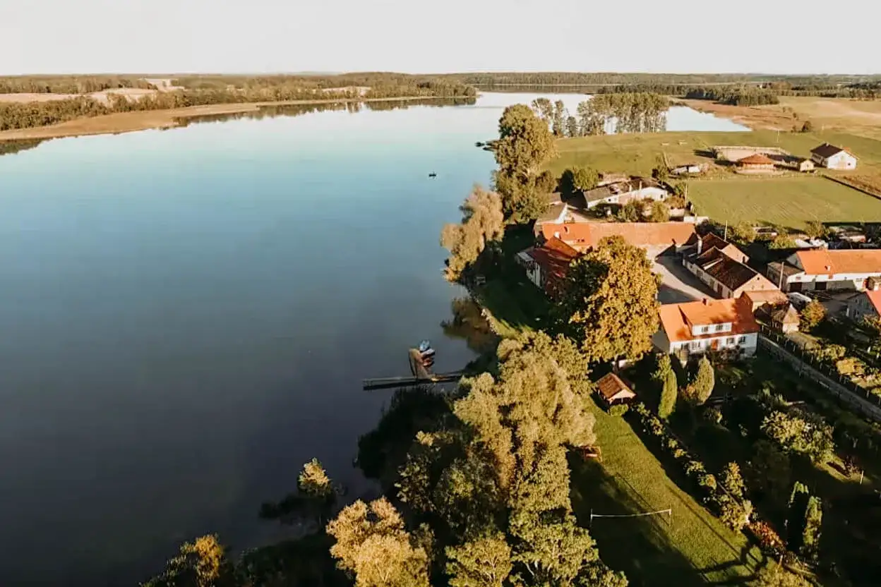 Spokojne jezioro Dejguny otoczone zielenią i domami z czerwonymi dachami. W oddali widać las.