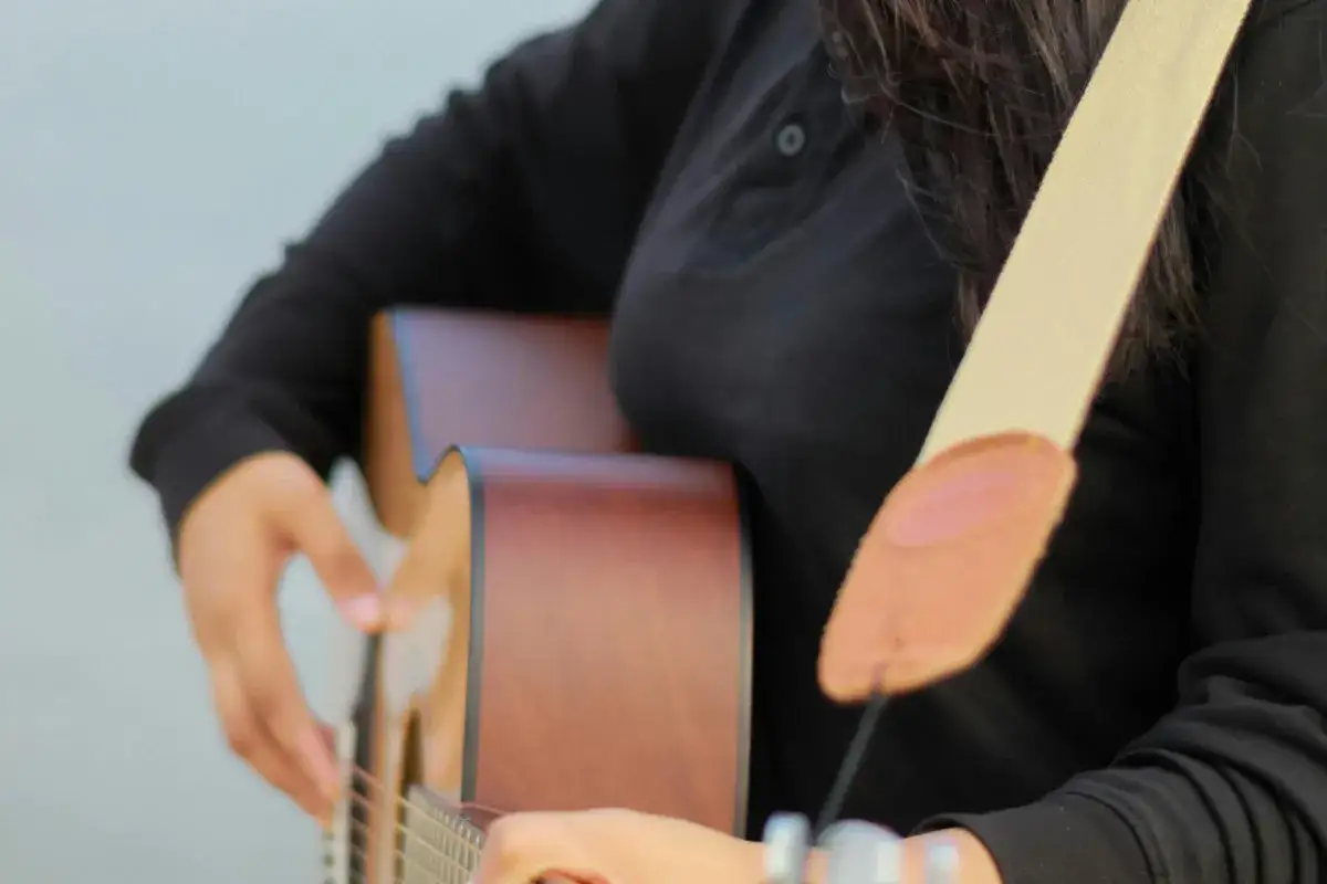 Osoba grająca na gitarze akustycznej, z beżowym paskiem przechodzącym przez ramię. Pokazuje, jak zaczepić pasek do gitary, by wygodnie grać.