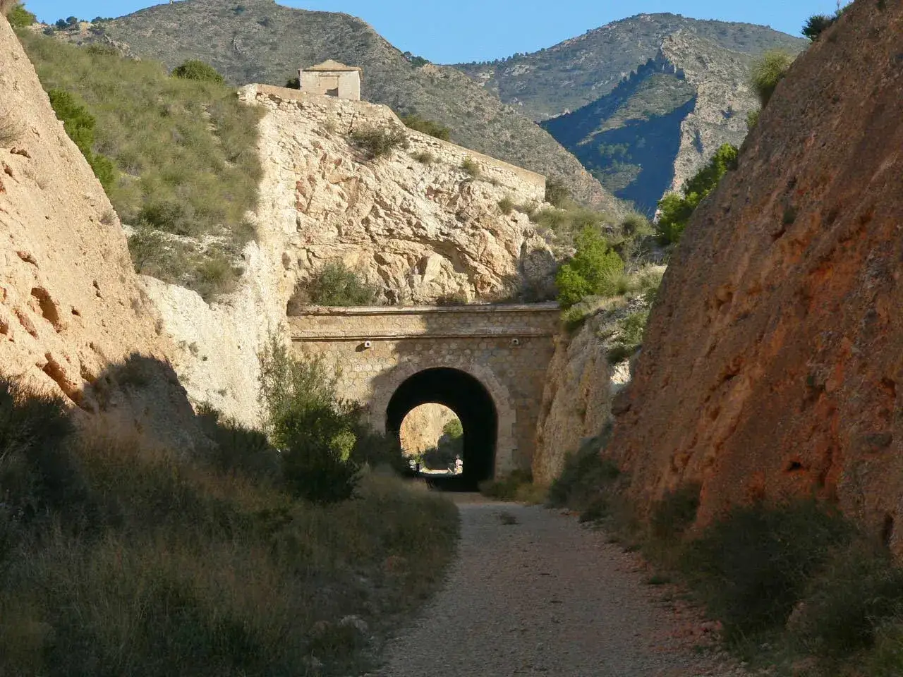 Camino de grava hacia un t&uacute;nel de piedra en la v&iacute;a verde de Alicante, rodeado de laderas rocosas y vegetaci&oacute;n.