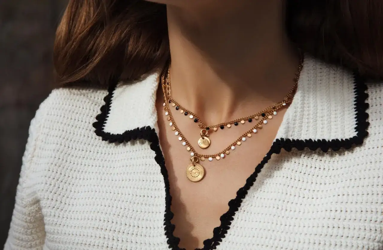 Elegancka biała narzutka z czarnym wykończeniem i złotymi naszyjnikami. Idealna moda po 50-tce stylizacje.