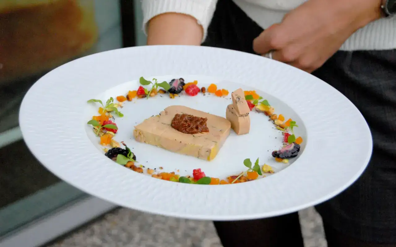 Portion foie gras par personne, joliment d&eacute;cor&eacute;e de fruits rouges, d'agrumes et de pistaches sur une assiette blanche.
