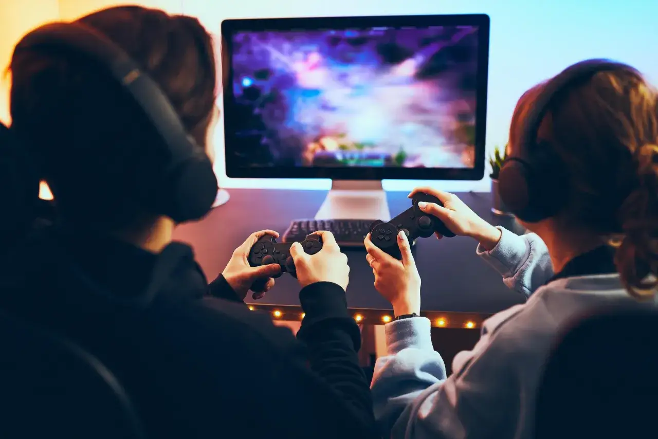 Zwei Personen mit Kopfhörern und Controllern vor einem leuchtenden Bildschirm. Sie sind vertieft in ihr Spiel, der Computer sucht nach neuen Herausforderungen.