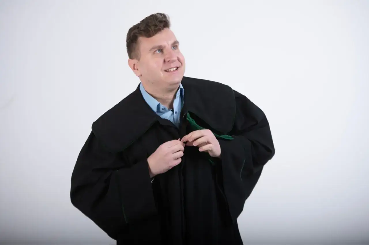Mężczyzna w todze prawniczej zapina płaszcz na tle białej ściany.