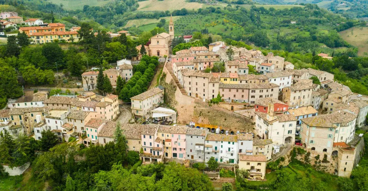 Borgo collinare nelle Marche, con case colorate e tetti in cotto, circondato da verde. Un luogo dove si respira la tradizione delle olive delle Marche.