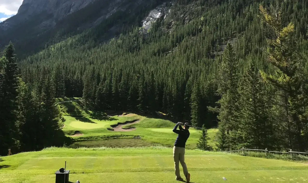En golfare svingar klubban på en bana omgiven av en tät granskog och höga berg. En dröm för golf norge.