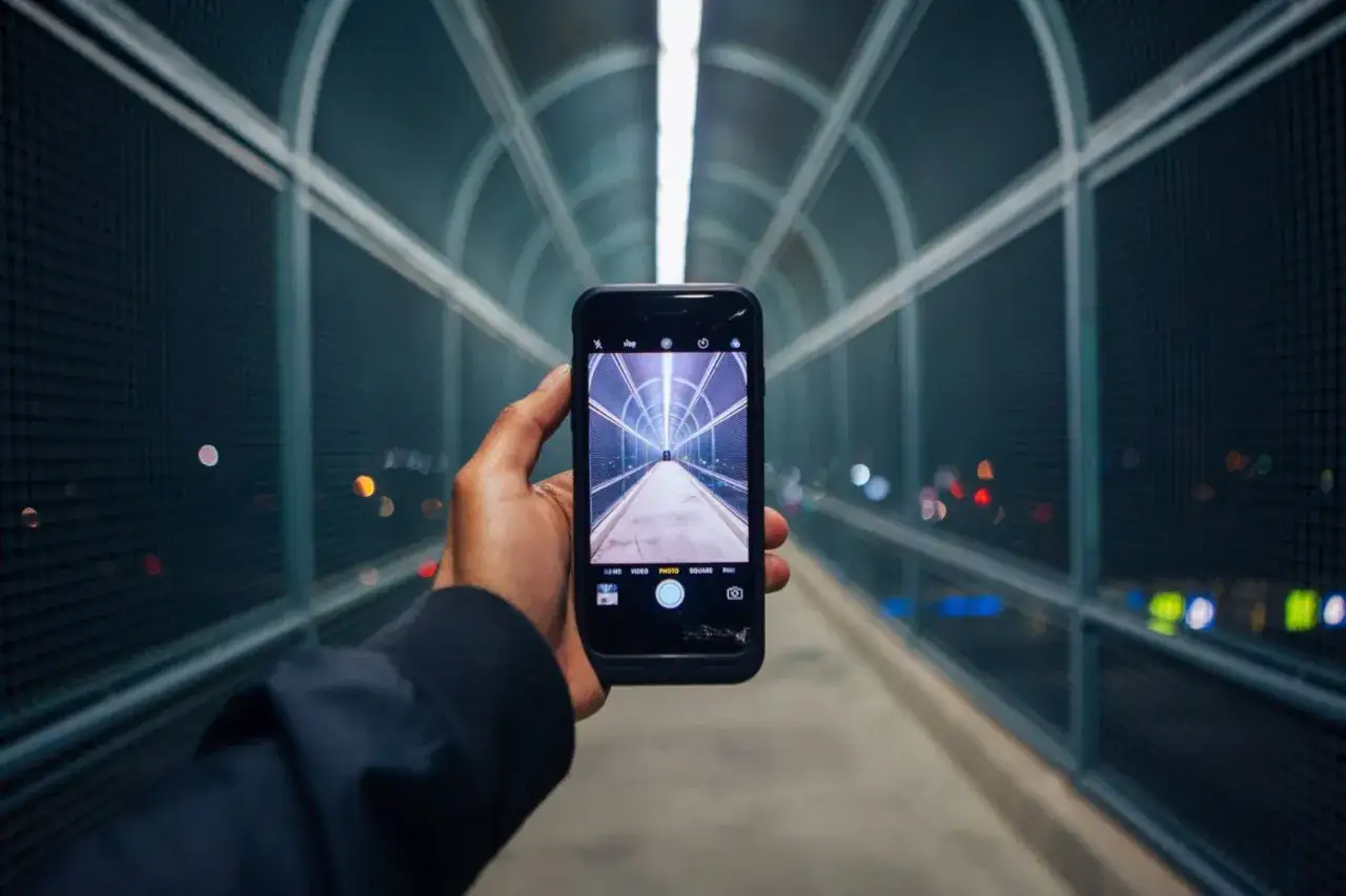 Dłoń trzyma smartfon, kt&oacute;rego aparat w telefonie rejestruje futurystyczny tunel z oświetleniem.