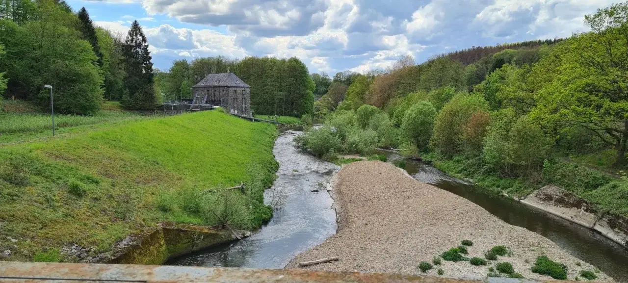 Ein malerischer Fluss schl&auml;ngelt sich durch eine gr&uuml;ne Landschaft, vorbei an einem alten Steingeb&auml;ude, das an das Schloss Ehreshoven erinnert.