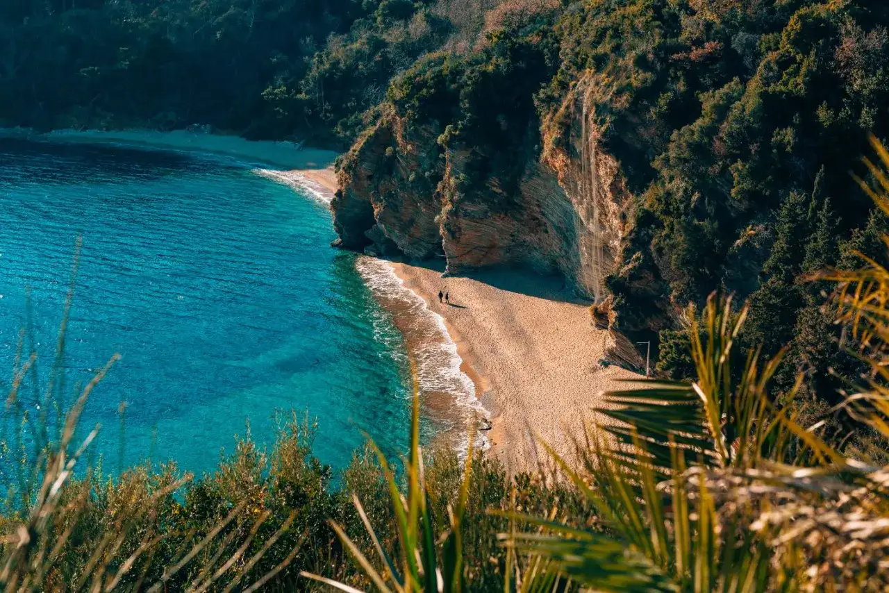 Dwie osoby spacerują po piaszczystej plaży otoczonej klifami, z turkusową wodą. To jedna z najpiękniejszych plaż Czarnogóry.