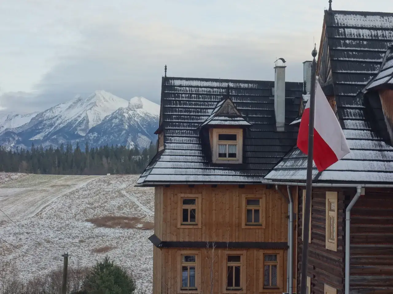 Drewniane domy w Bukowinie Tatrzańskiej, rynek z widokiem na ośnieżone góry. Polska flaga powiewa na wietrze.