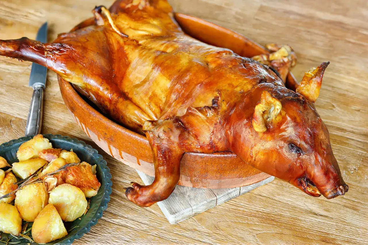 A golden-brown, whole roasted spanish lechon sits in a rustic clay dish, accompanied by a side of crispy roasted potatoes.