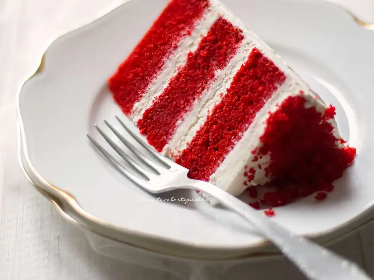 Una fetta di torta Red Velvet, con strati di pan di Spagna rosso intenso e crema bianca, pronta per essere gustata. La ricetta perfetta per un dolce indimenticabile.