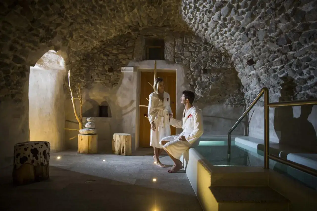Coppia rilassata in accappatoio, godendosi l'atmosfera suggestiva delle Caderzone Terme, con vasca d'acqua calda e pareti in pietra.
