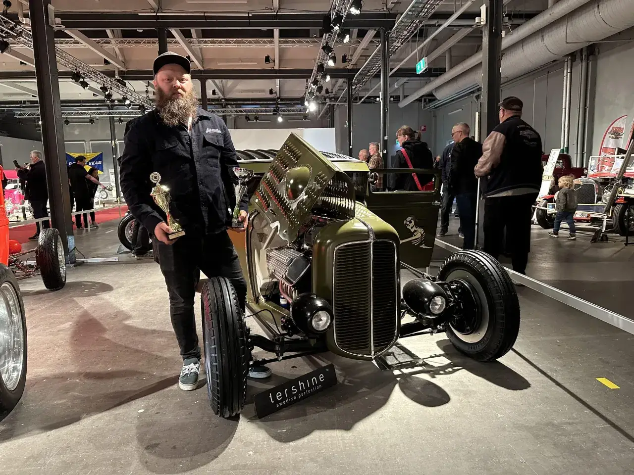 En stolt svensk hot rod byggare med pokaler bredvid sin gröna custombil.