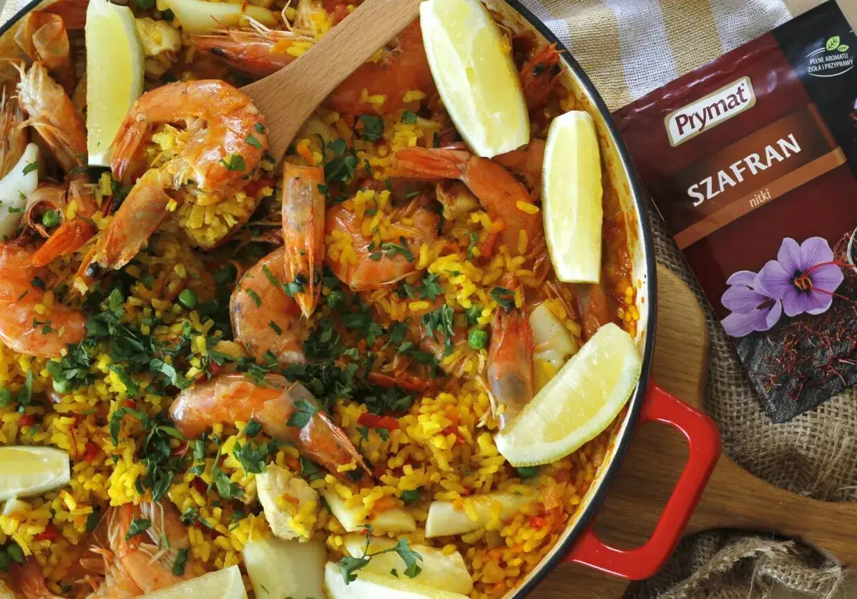 Pyszna paella z kurczakiem i krewetkami, ozdobiona cytryną i natką pietruszki. Obok paczka szafranu, kluczowego składnika tej potrawy.