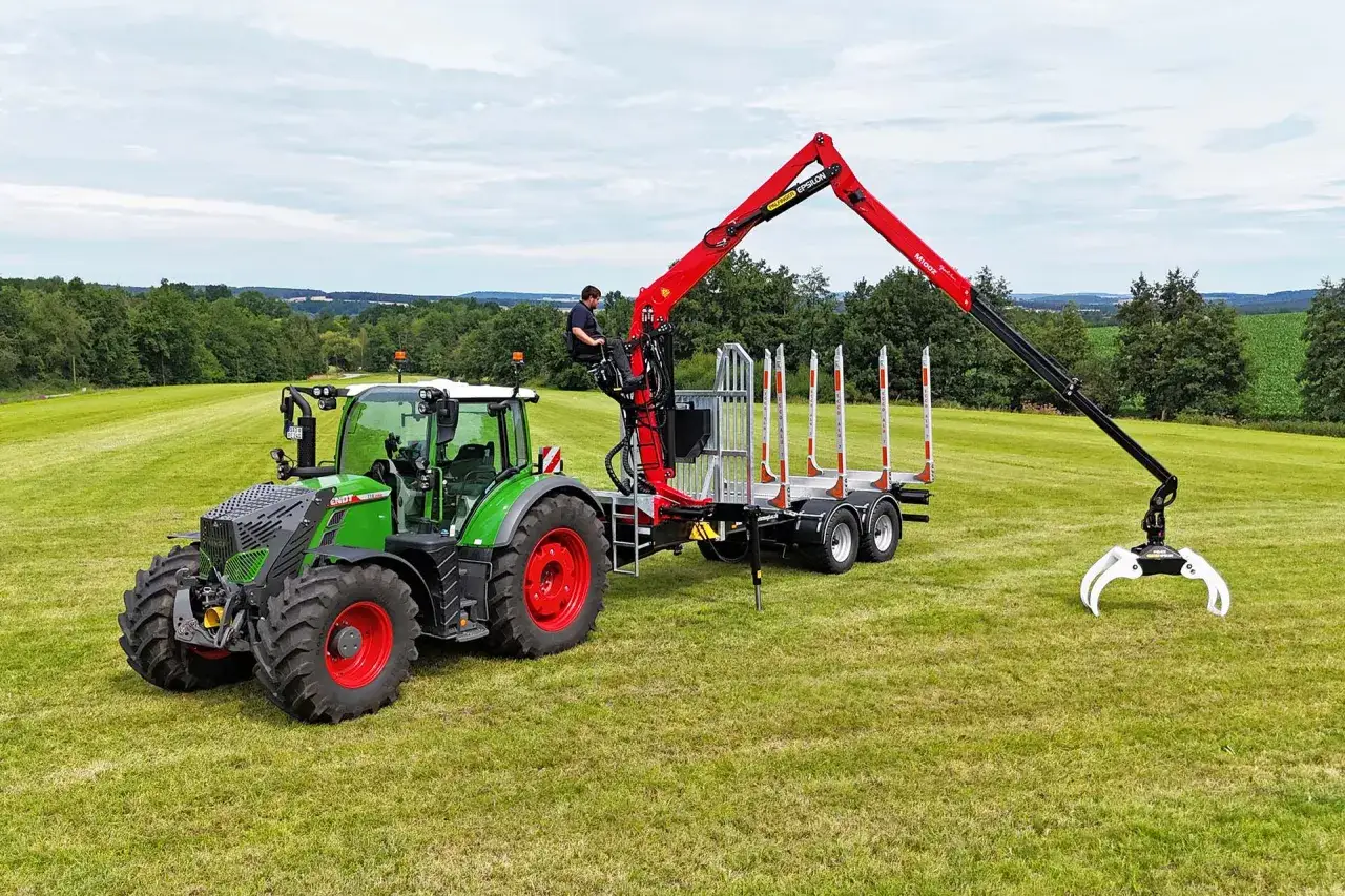 Gr&uuml;ner Traktor mit angekoppeltem Anh&auml;nger und einem roten Ladekran, bereit f&uuml;r den Holzeinsatz im Wald.