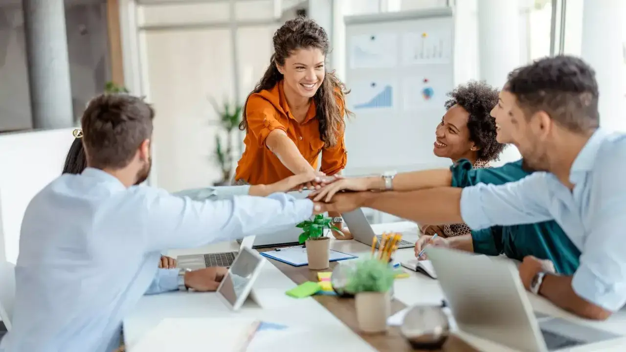 A diverse team stacks hands, smiling, embodying how to create psychological safety through unity and shared purpose.