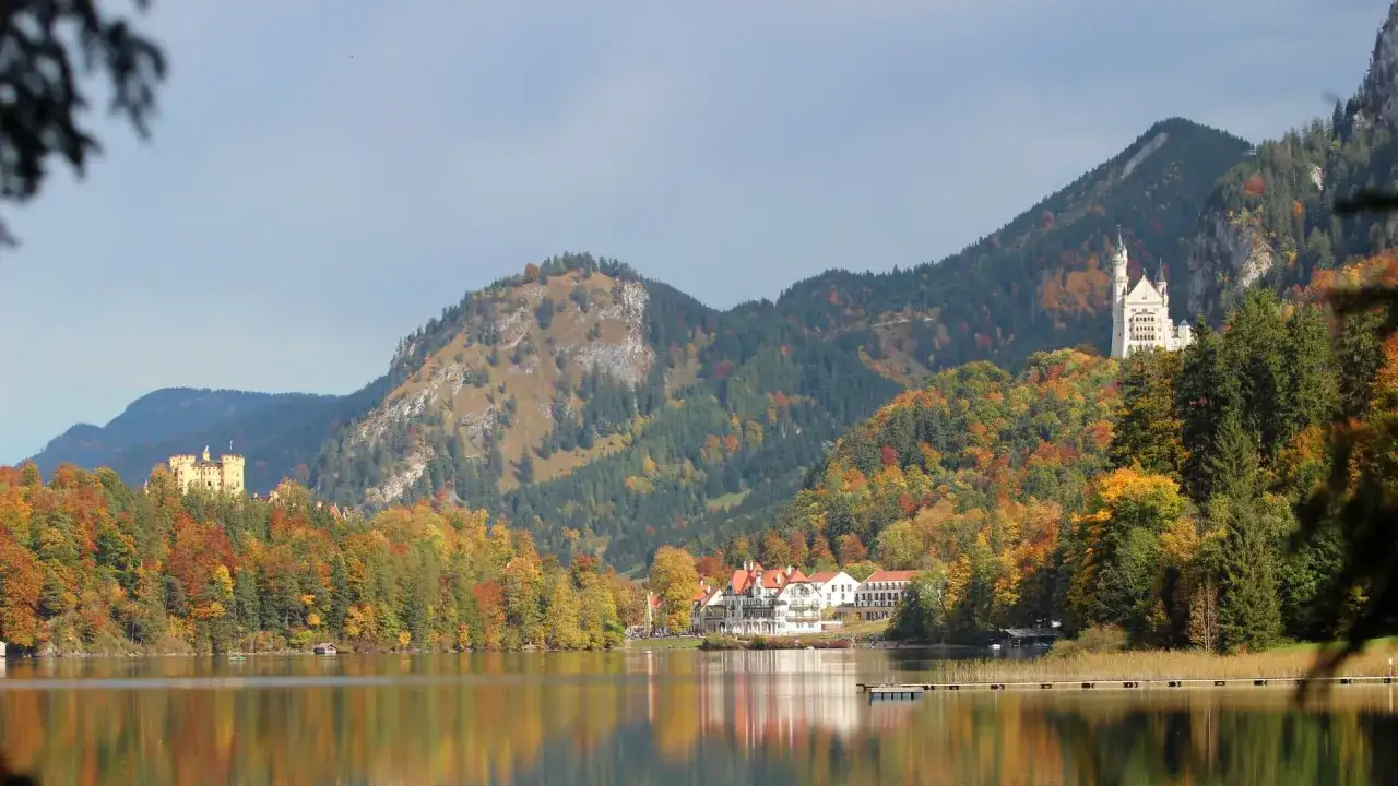 Herbstliche **Unternehmungen in Bayern**: Zwei Schlösser thronen auf bewaldeten Hügeln über einem spiegelglatten See.