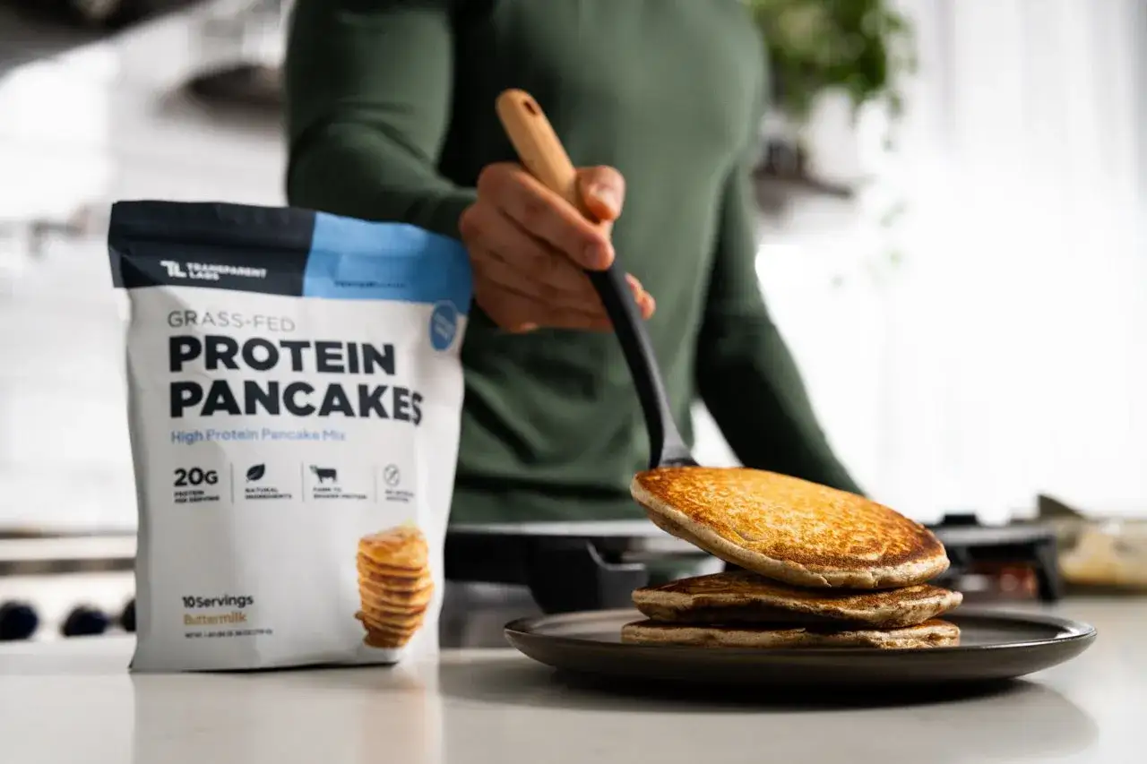Frisch zubereitete, goldbraune **high protein pancakes** werden auf einen Teller gelegt. Daneben steht die Packung der Protein Pancake Mix.