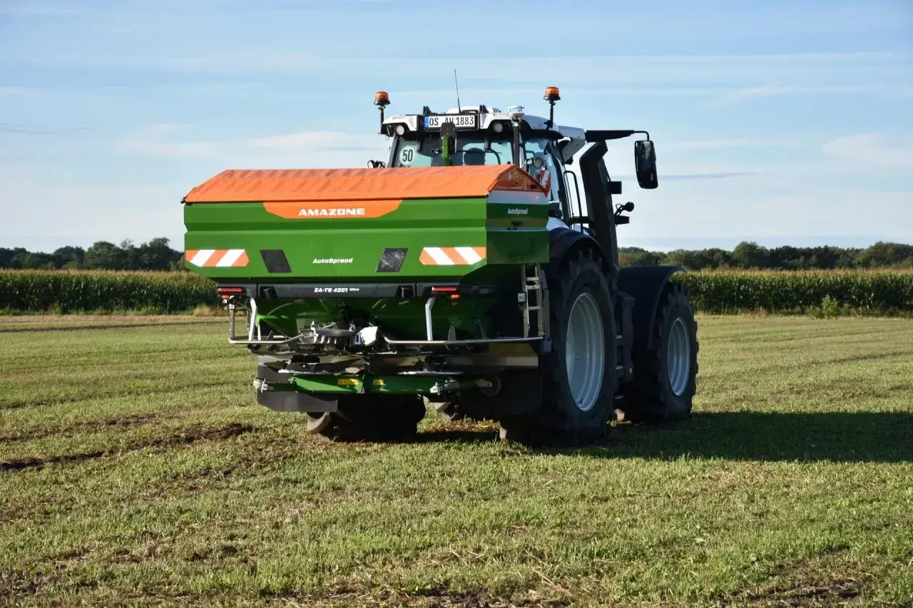 Traktor z rozsiewaczem AMAZONE wykonuje nawożenie jęczmienia ozimego na polu.