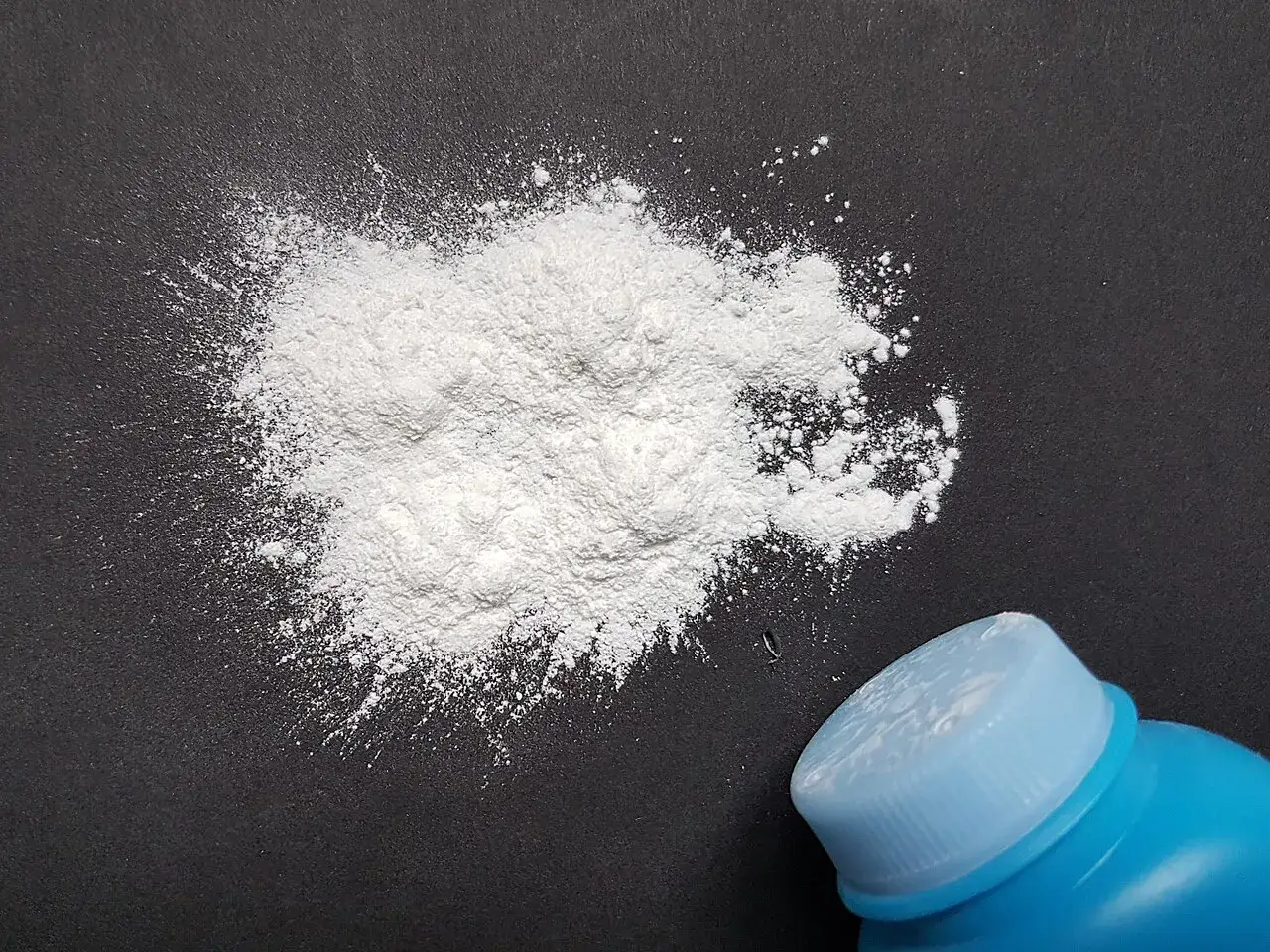 Una nuvola di borotalco bianco su sfondo scuro, vicino a un tappo blu. Attenzione: il borotalco &egrave; cancerogeno.