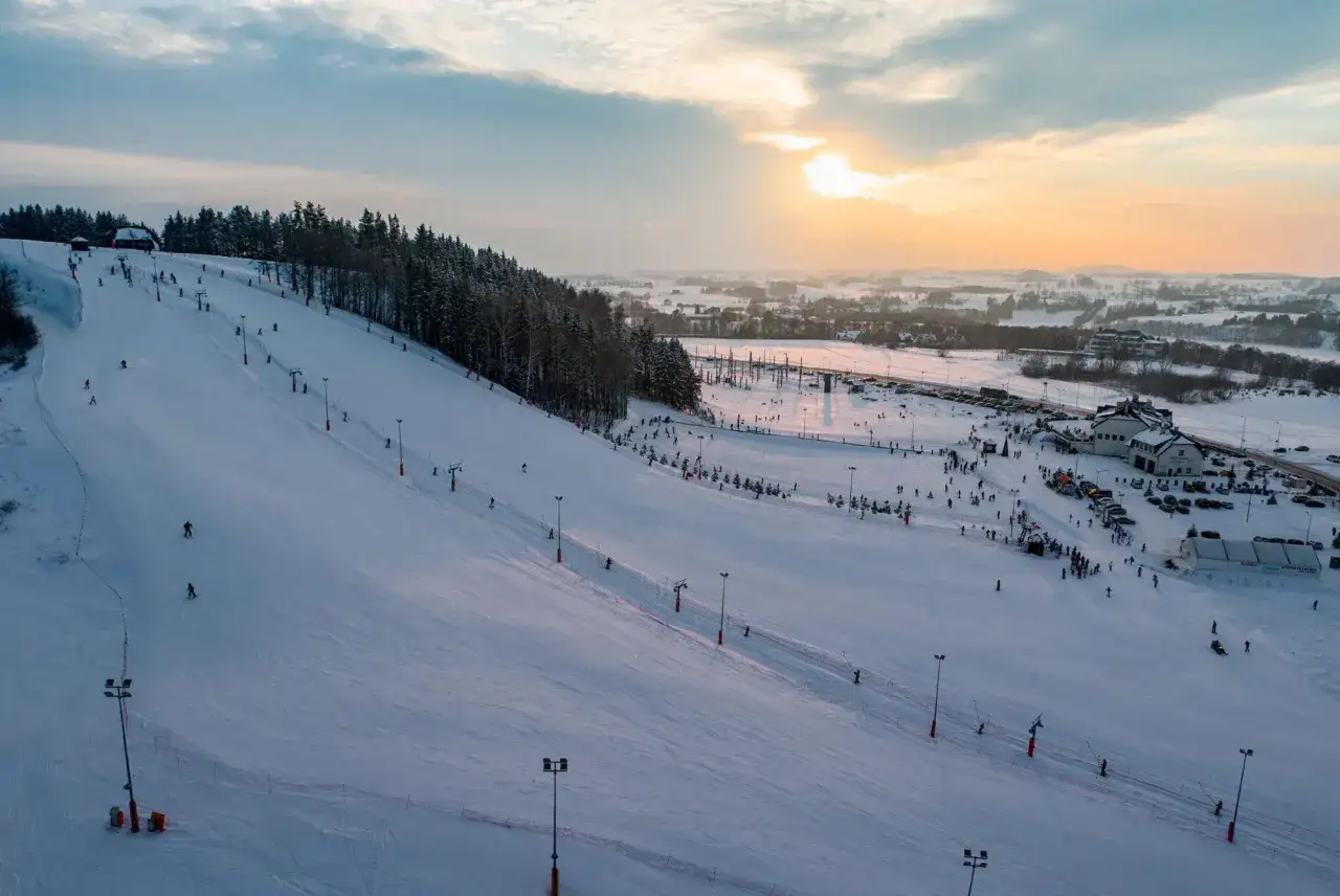 Ośnieżone stoki narciarskie Hańcza Ski Resort tętnią życiem. Narciarze szusują, a w tle widać budynki i parkingi.