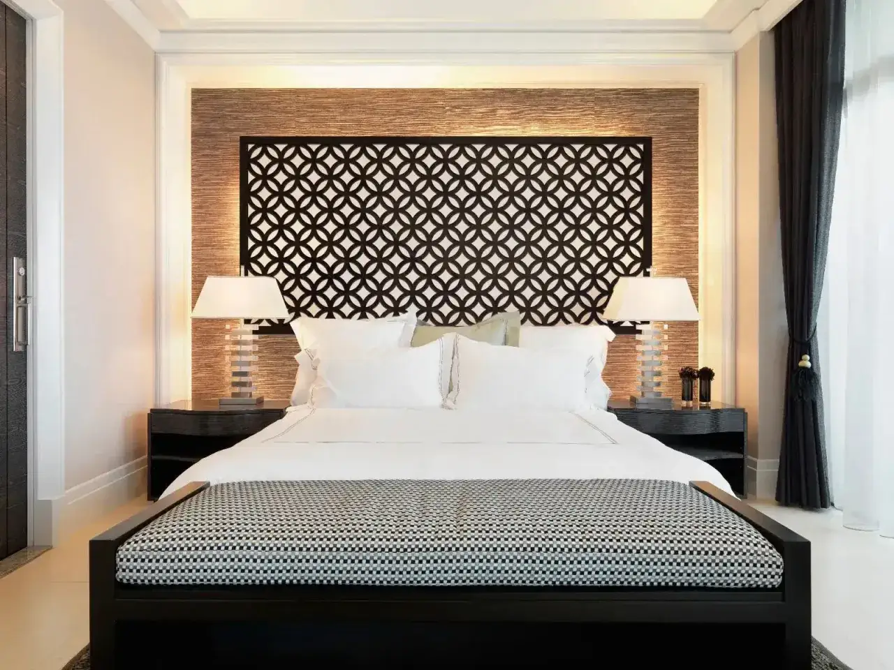 Elegante dormitorio de matrimonio sin cabecero, con cama blanca, mesitas negras y l&aacute;mparas modernas. El cabecero es un panel decorativo con un patr&oacute;n geom&eacute;trico.