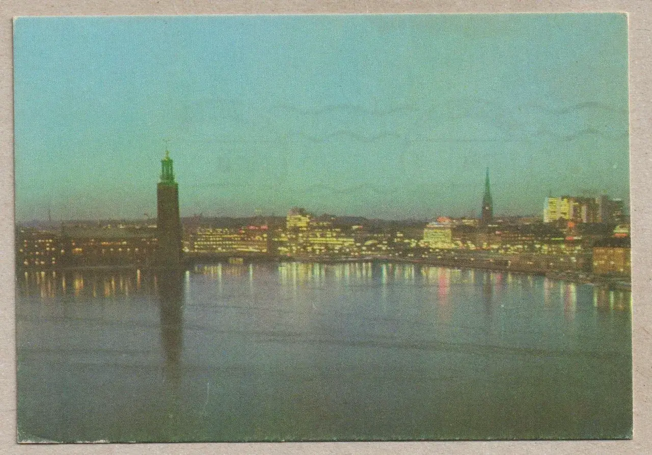 Stockholms stadshus reser sig mot en bl&aring; kv&auml;llshimmel, med upplysta byggnader l&auml;ngs vattnet som speglar sig i det lugna vattnet.
