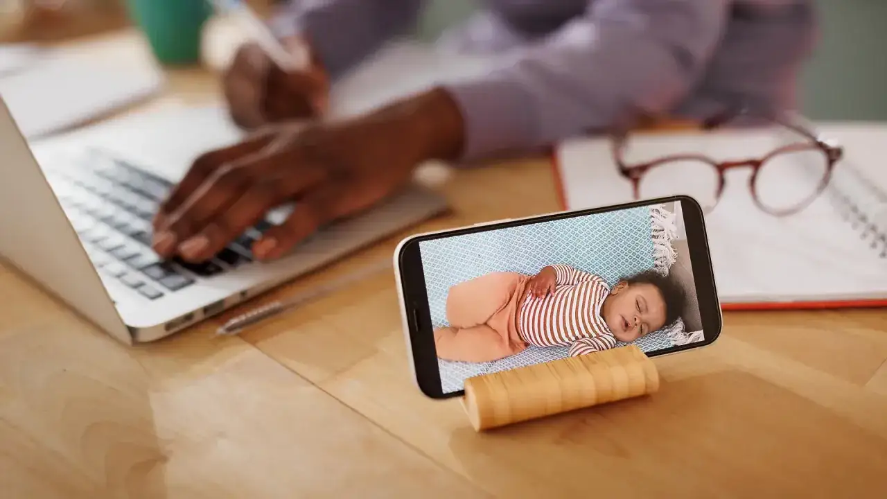 Ein Handy dient als Babyphone und zeigt ein schlafendes Baby. Im Hintergrund arbeitet jemand am Laptop.