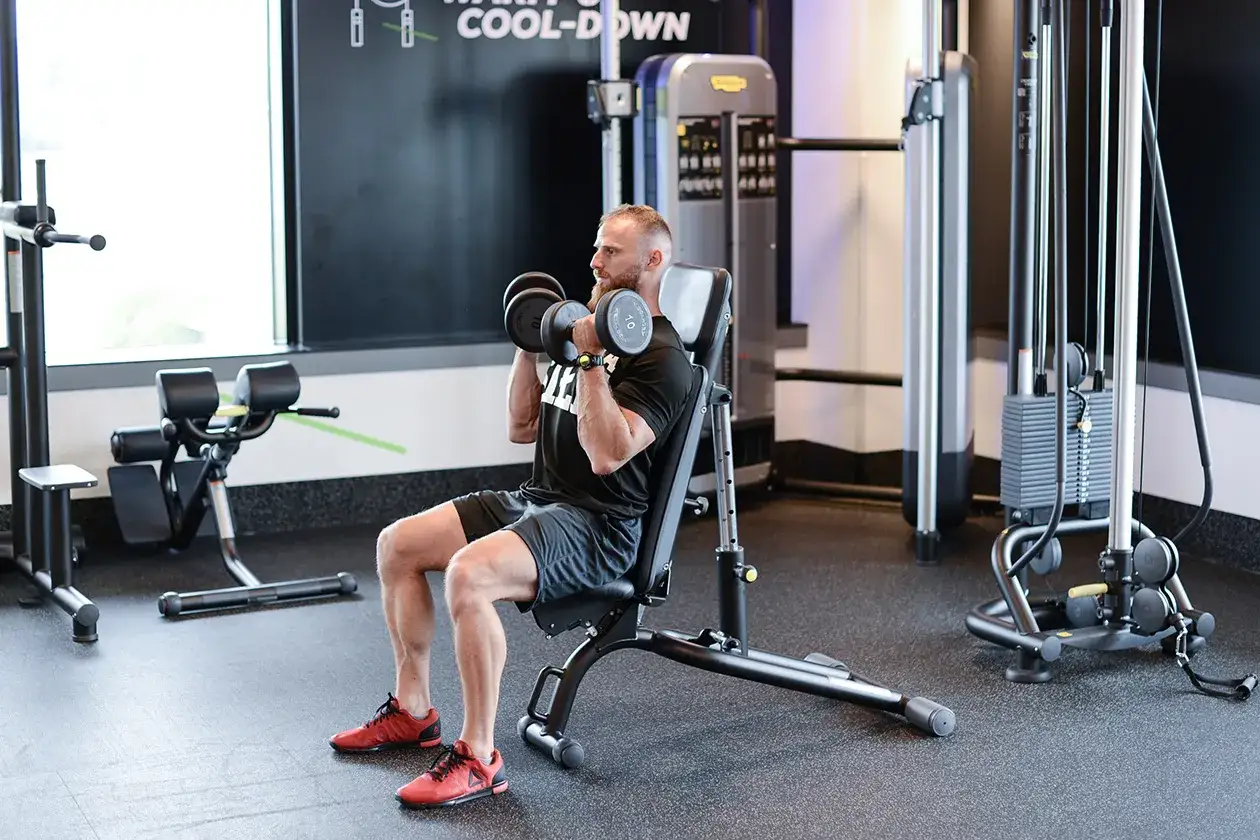 Mężczyzna ćwiczy z hantlami na ławce, wykonując ćwiczenie na biceps. Arnoldki stojąc to jego cel.