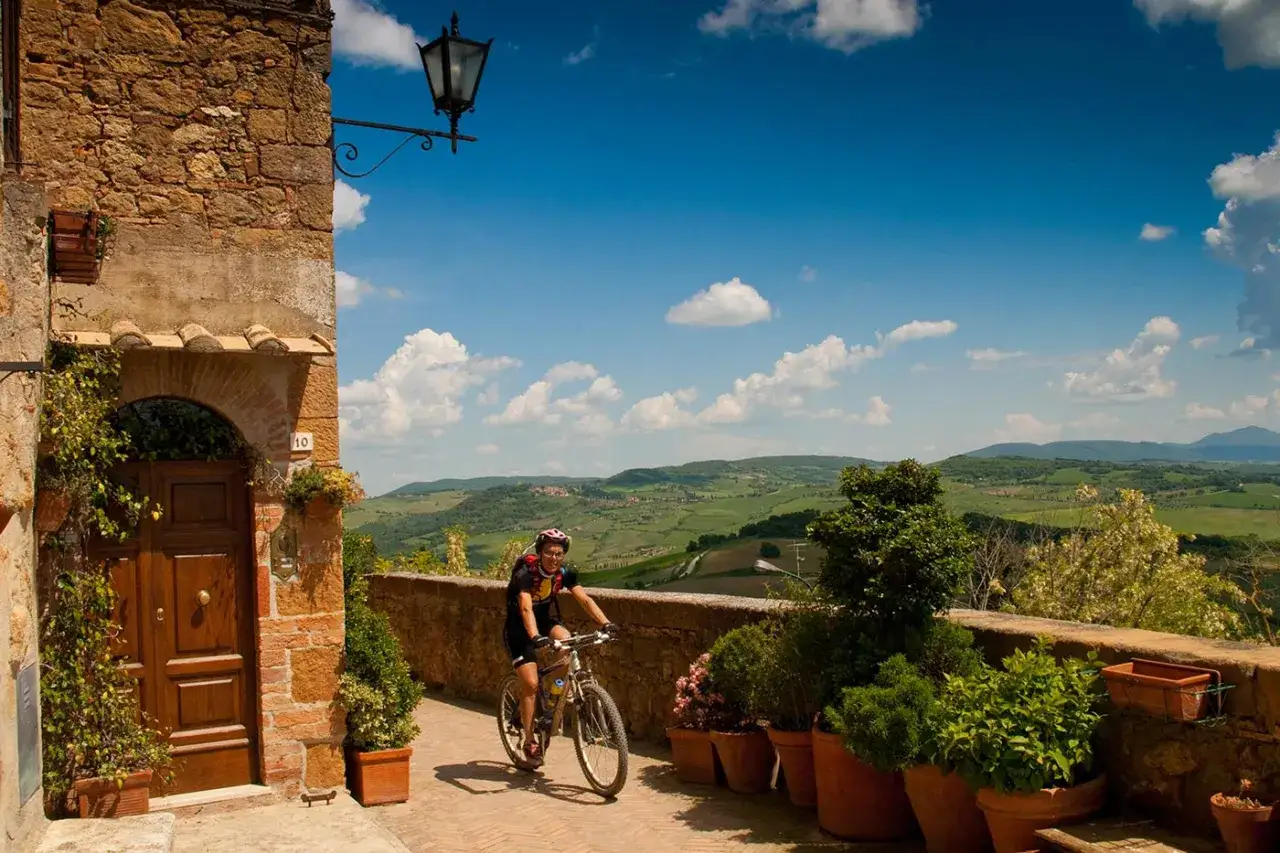 Donna in bici esplora il Val d'Orcia, tra antiche mura e colline verdi sotto un cielo azzurro.