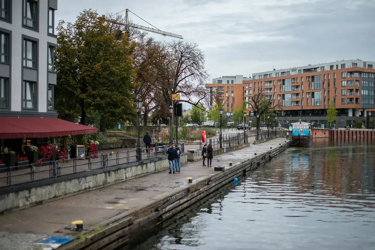 Spacerowicze na nabrzeżu, budynki mieszkalne i ł&oacute;dź z napisem 